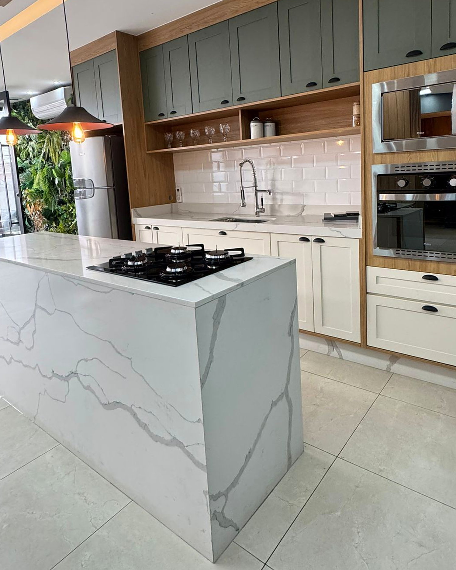 Cozinha planejada com ilha em quartzo, cooktop embutido, armários verde-sálvia e torre de fornos em inox