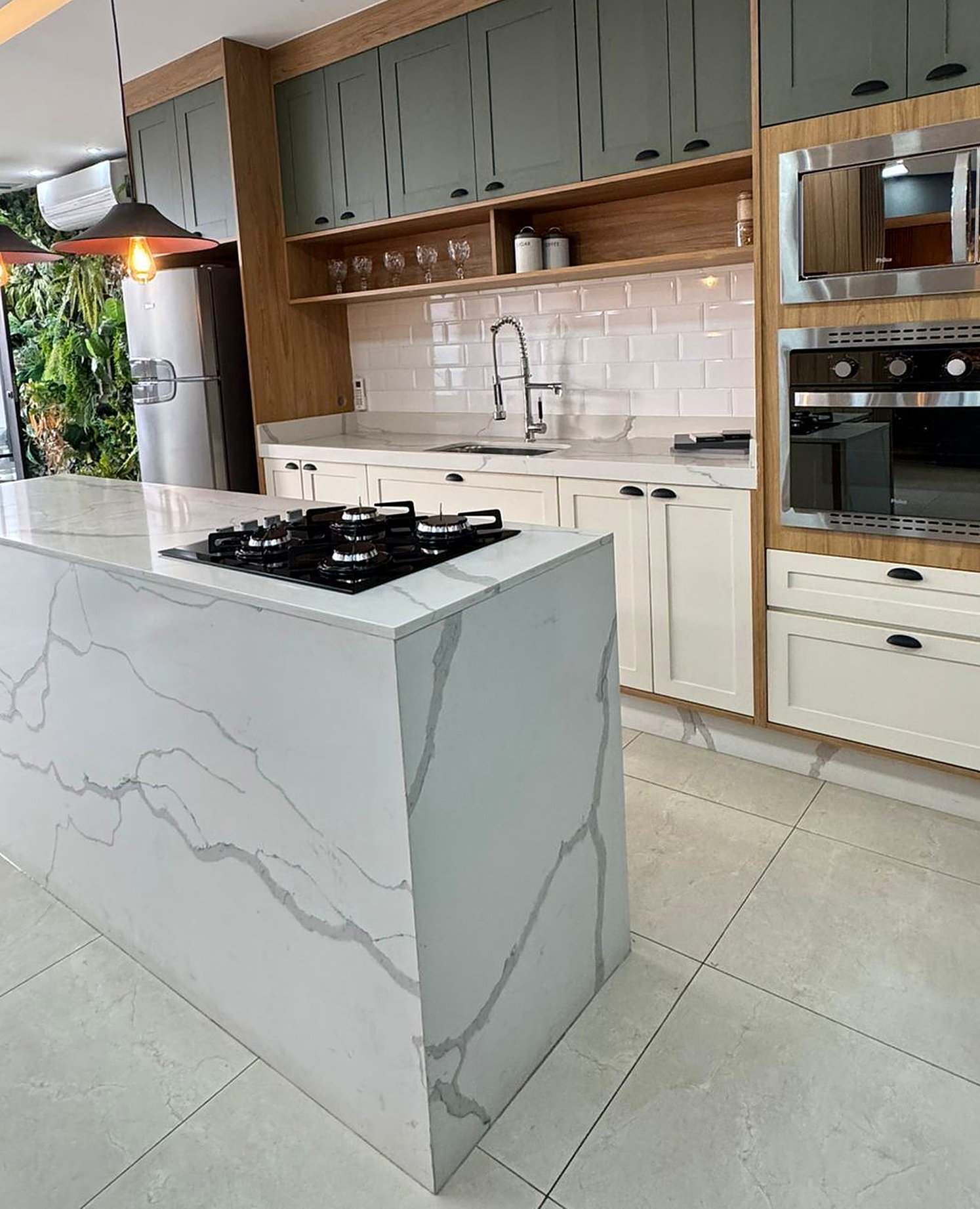 Ilha de cozinha planejada em quartzo branco com cooktop embutido, armários em verde-sálvia e nichos amadeirados ao fundo — projeto da 2L Concept